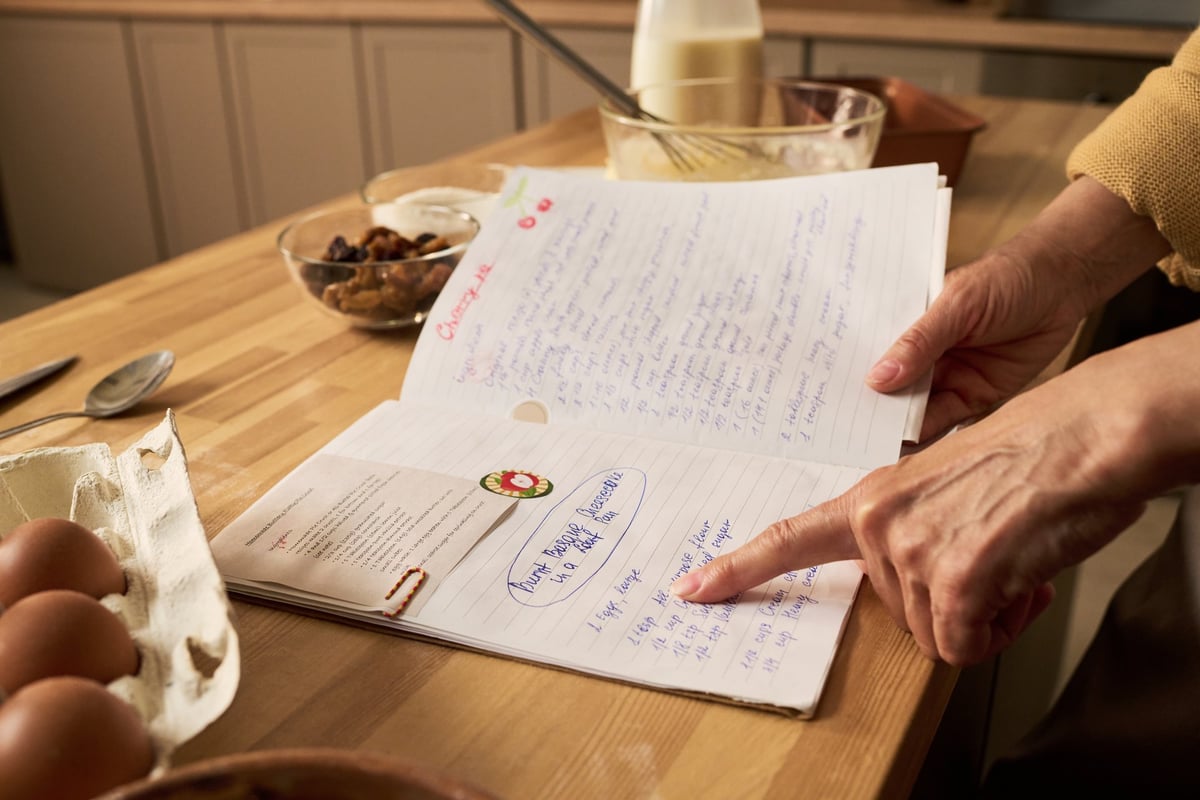 A woman writing notes in a recipe notebook beside eggs and milk on a kitchen counter