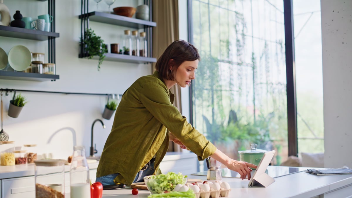 A woman following a recipe on a tablet by a bright kitchen window