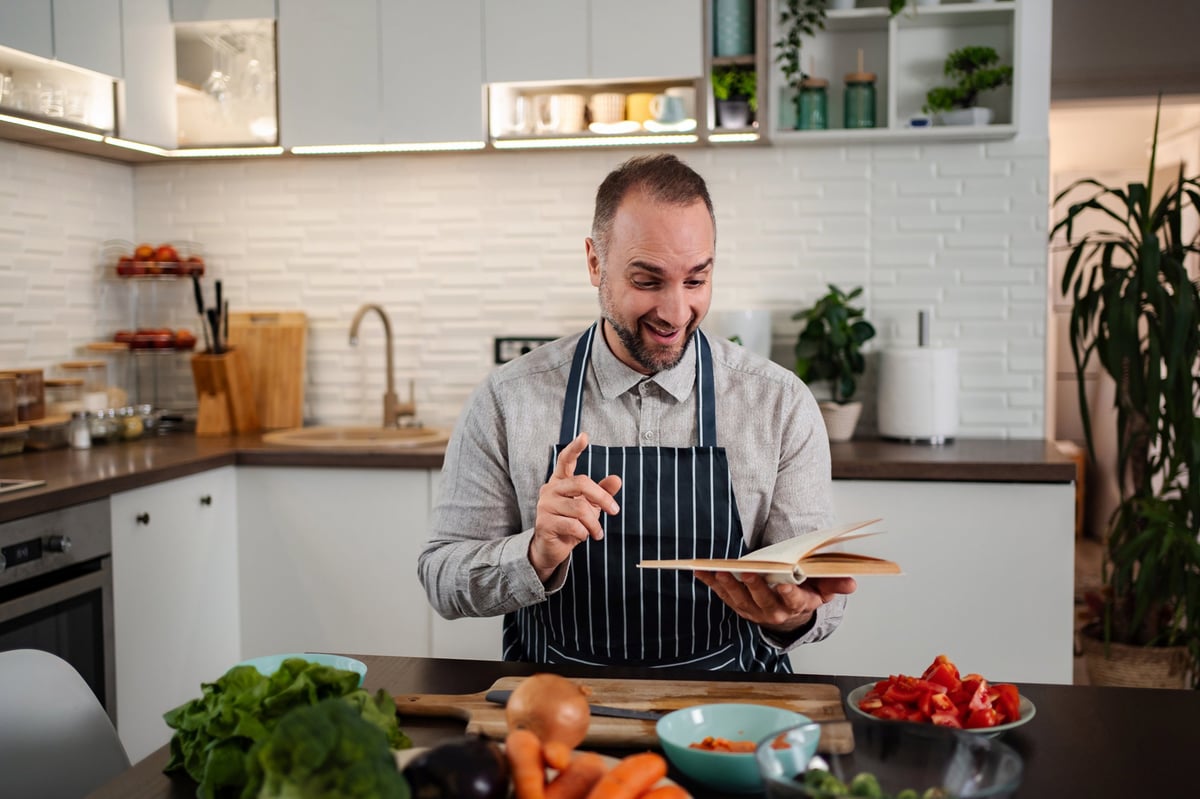 A man reading a cookbook while preparing fresh vegetables